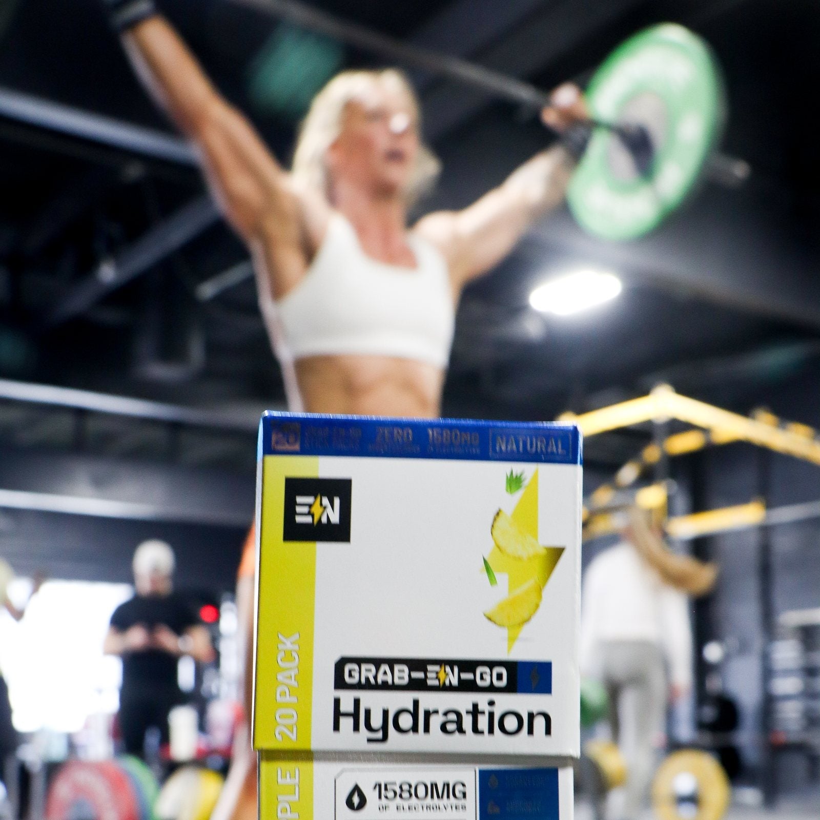 Engineered Nutrition Grab-EN-Go hydration stick pack box on a gym platform while a CrossFit athlete presses a barbell overhead behind it