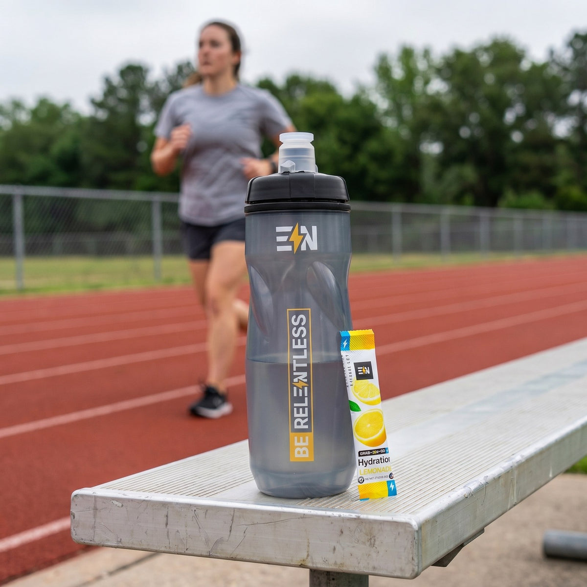Engineered Nutrition squeeze bottle and Lemonade hydration stick on a trackside bench while a runner jogs a lap behind them