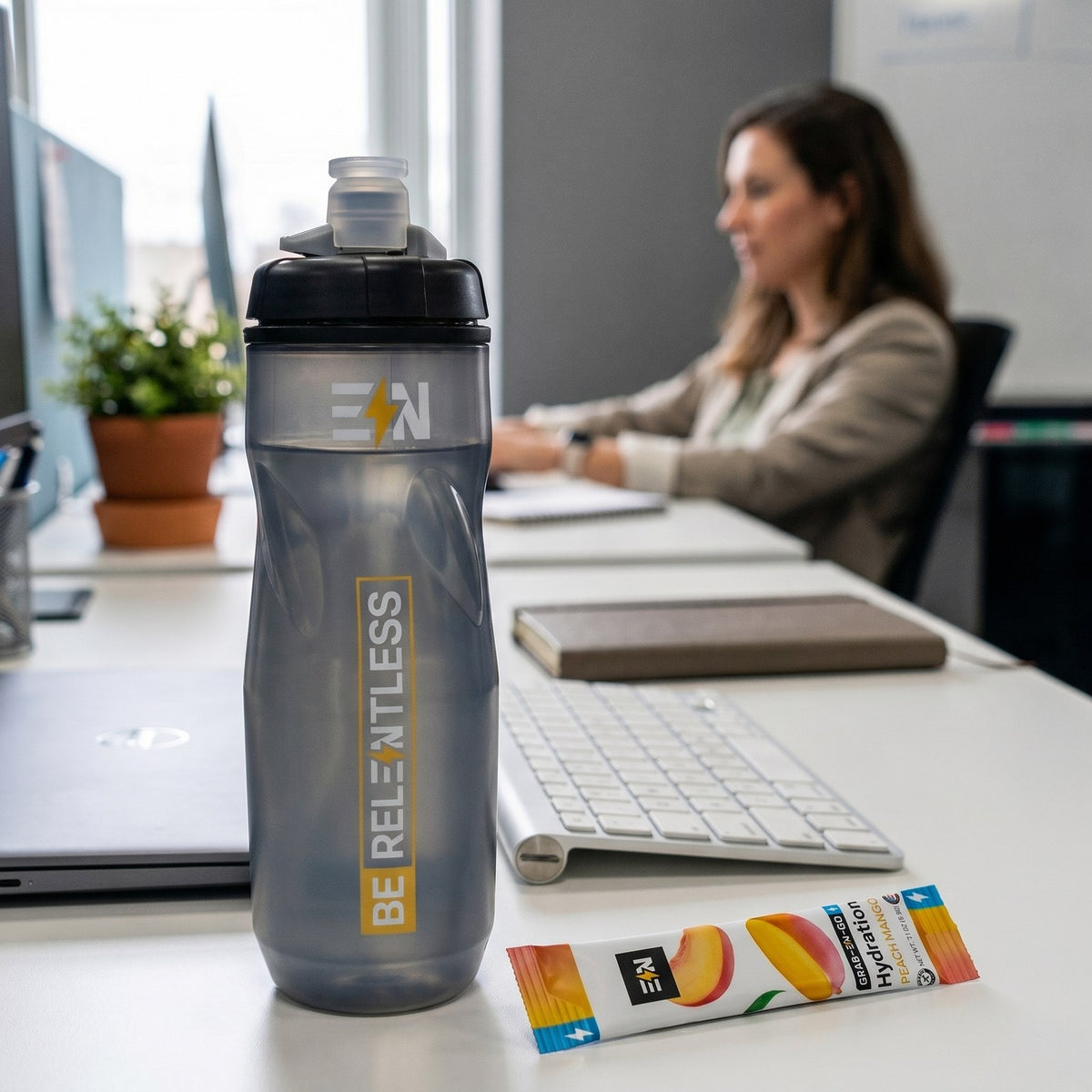 Engineered Nutrition squeeze bottle and Peach Mango hydration stick on an office desk while a woman works behind them
